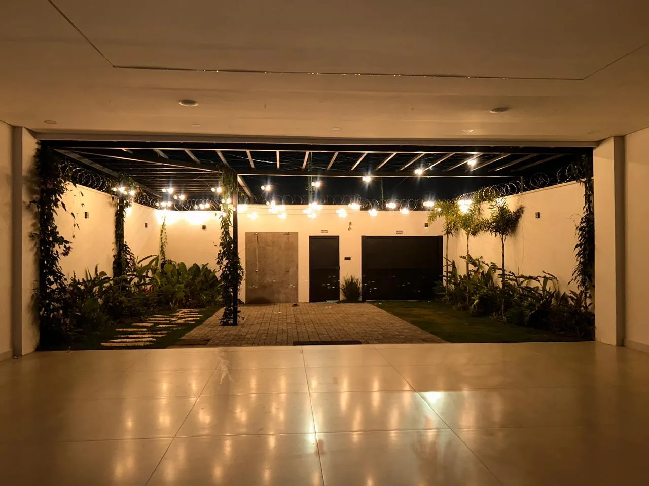 Salão de festas iluminado à noite, com abertura para o jardim e pergolado com luzes, em Uberlândia/MG